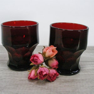 May include: Two dark red glass tumblers with a faceted design. The tumblers are sitting on a wooden surface with a small bouquet of pink roses in front of them.