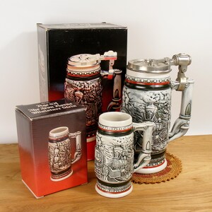 May include: Three ceramic beer steins with a train theme. The largest stein has a metal lid and handle. The smallest stein is in a box that says "Age of the Iron Horse Miniature Stein".