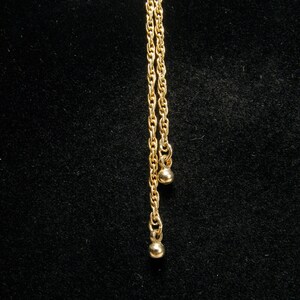 May include: A gold-coloured necklace featuring a bow-shaped pendant and two hanging chains. Each chain has a small, round charm at the end. The necklace is set against a black background.