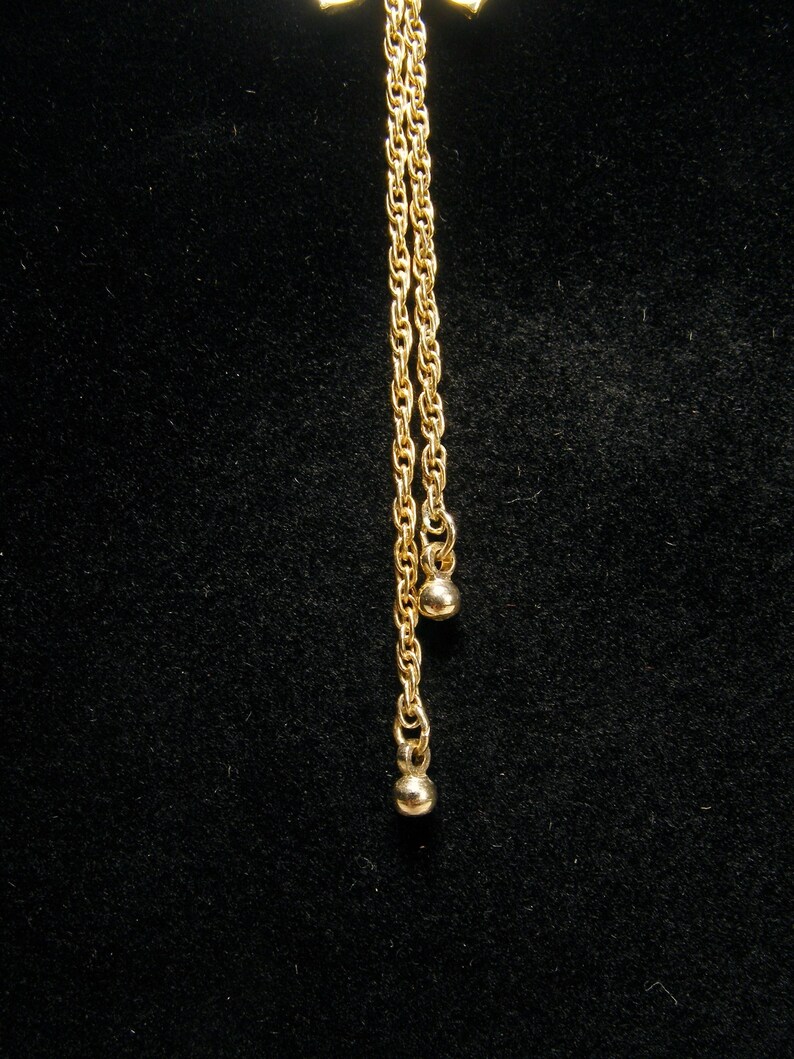 May include: A gold-coloured necklace featuring a bow-shaped pendant and two hanging chains. Each chain has a small, round charm at the end. The necklace is set against a black background.