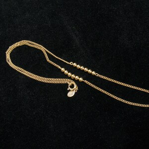 May include: A delicate gold necklace with a chain and small, round beads. The necklace has a lobster clasp closure. The jewelry is displayed on a black surface.