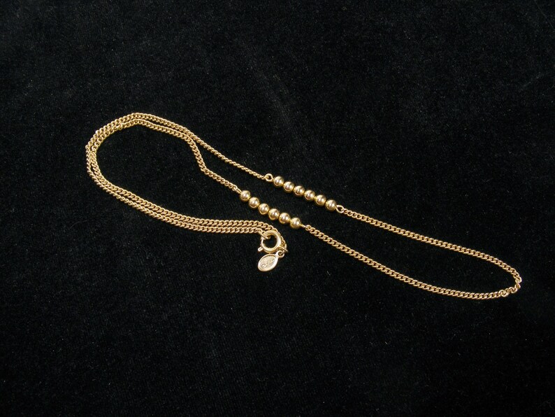 May include: A delicate gold necklace with a chain and small, round beads. The necklace has a lobster clasp closure. The jewelry is displayed on a black surface.