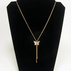 May include: Gold-coloured butterfly pendant necklace. The butterfly pendant is decorated with small, sparkling stones. The necklace has a delicate chain and a longer, dangling chain with small, round accents. It is displayed on a black velvet jewellery stand.