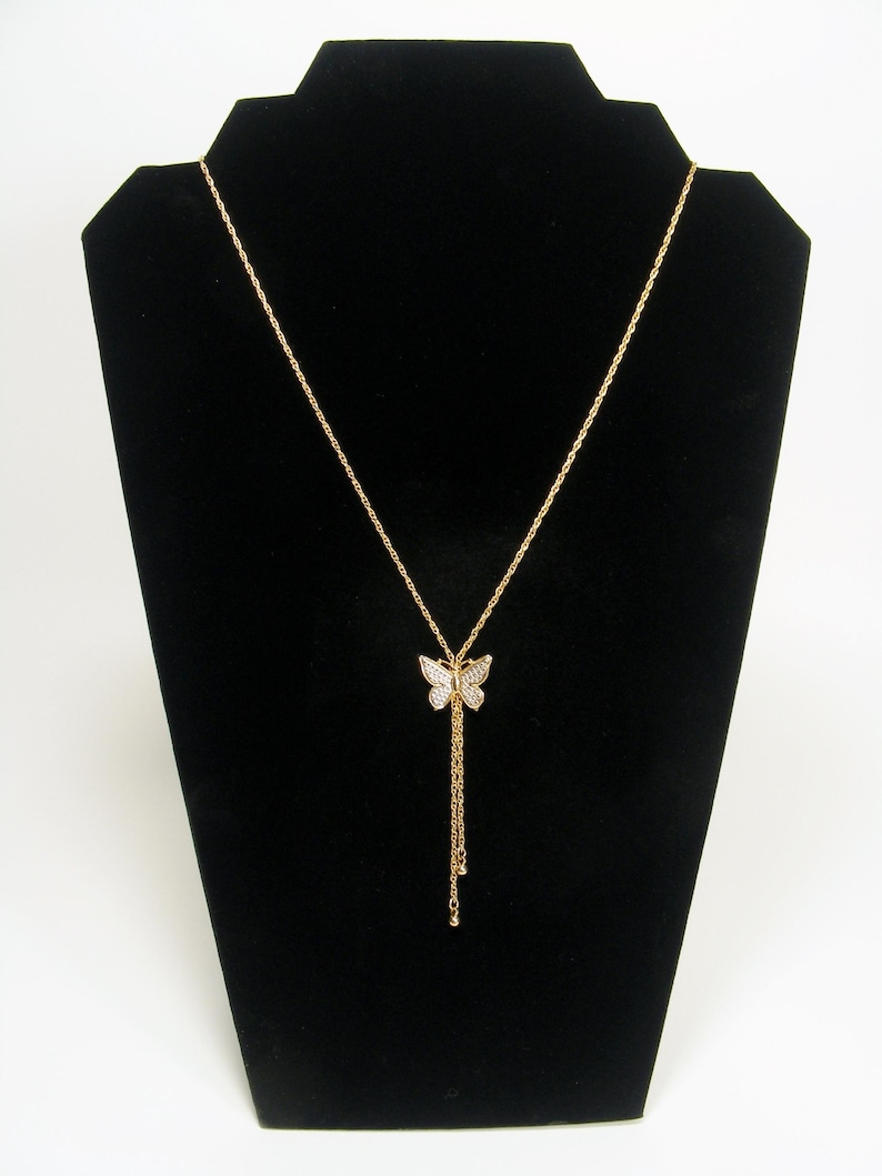 May include: Gold-coloured butterfly pendant necklace. The butterfly pendant is decorated with small, sparkling stones. The necklace has a delicate chain and a longer, dangling chain with small, round accents. It is displayed on a black velvet jewellery stand.