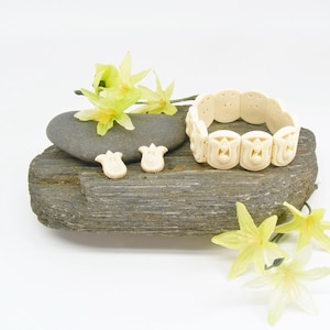 May include: An ivory-coloured jewellery set, comprising a bracelet and two earrings, presented on a grey stone. The bracelet is made of carved, linked segments. The earrings share a similar design. Yellow flowers provide a splash of colour.