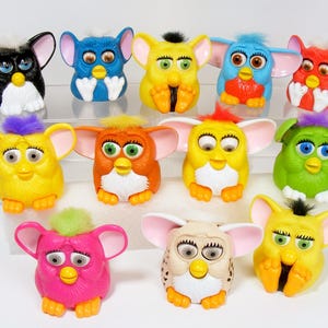 May include: A collection of colorful Furby toys. The toys have large ears, expressive eyes, and a variety of colors, including black, blue, yellow, red, green, orange, pink, and tan. Each Furby has a unique tuft of hair on its head.