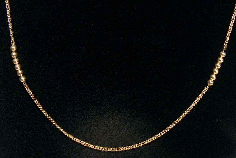 May include: A delicate gold necklace with a curb chain and small, round beads. The necklace is displayed against a black background, highlighting its simple elegance. The chain is approximately 45 cm long.