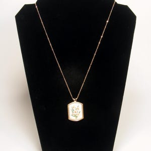 May include: A gold-coloured necklace with a rectangular pendant featuring a floral design. The pendant has a white background with a gold border. The necklace is displayed on a black velvet jewellery stand.