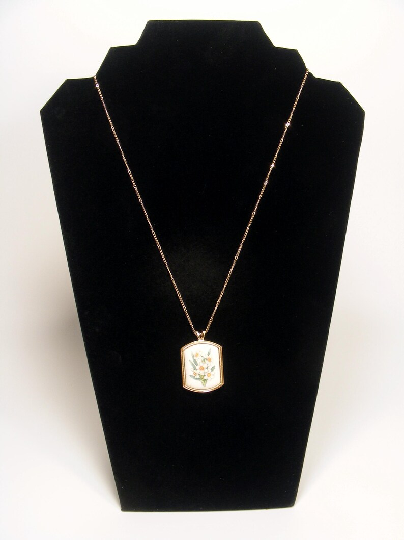 May include: A gold-coloured necklace with a rectangular pendant featuring a floral design. The pendant has a white background with a gold border. The necklace is displayed on a black velvet jewellery stand.