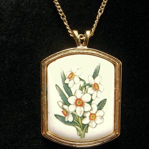 May include: A gold-coloured pendant necklace. The rectangular pendant displays a floral design of white daffodils with orange centres and green leaves on a white background. The chain is a delicate gold colour, suitable for everyday wear.