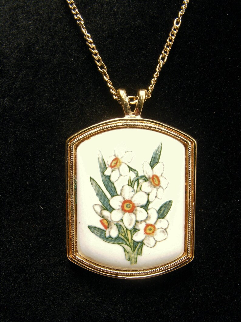 May include: A gold-coloured pendant necklace. The rectangular pendant displays a floral design of white daffodils with orange centres and green leaves on a white background. The chain is a delicate gold colour, suitable for everyday wear.