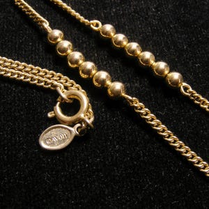 May include: A gold-coloured necklace with a fine chain and beaded details. The necklace has a lobster clasp and an oval tag with the "AVON" logo. The jewellery is set against a dark background.