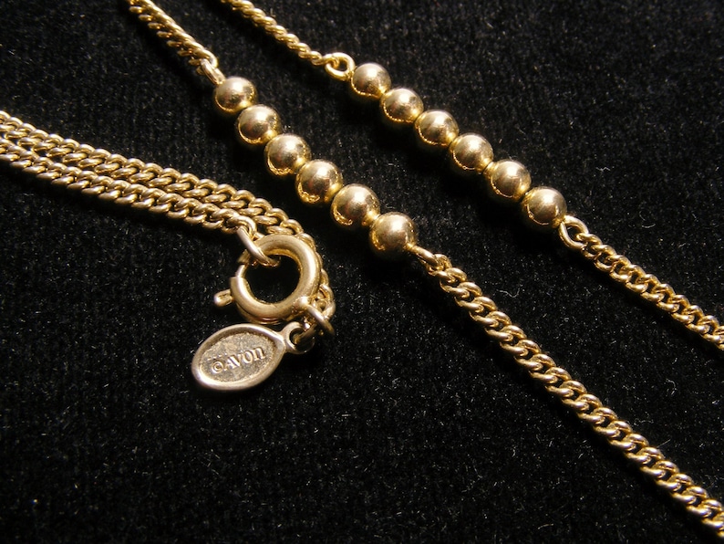 May include: A gold-coloured necklace with a fine chain and beaded details. The necklace has a lobster clasp and an oval tag with the "AVON" logo. The jewellery is set against a dark background.