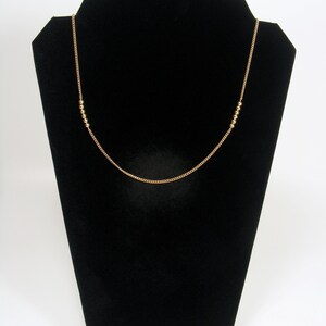 May include: A gold-coloured necklace on a black jewellery stand. The necklace has a fine chain with small, round beads spaced along it. The design is simple and elegant, suitable for everyday wear, measuring approximately 45 cm in length.