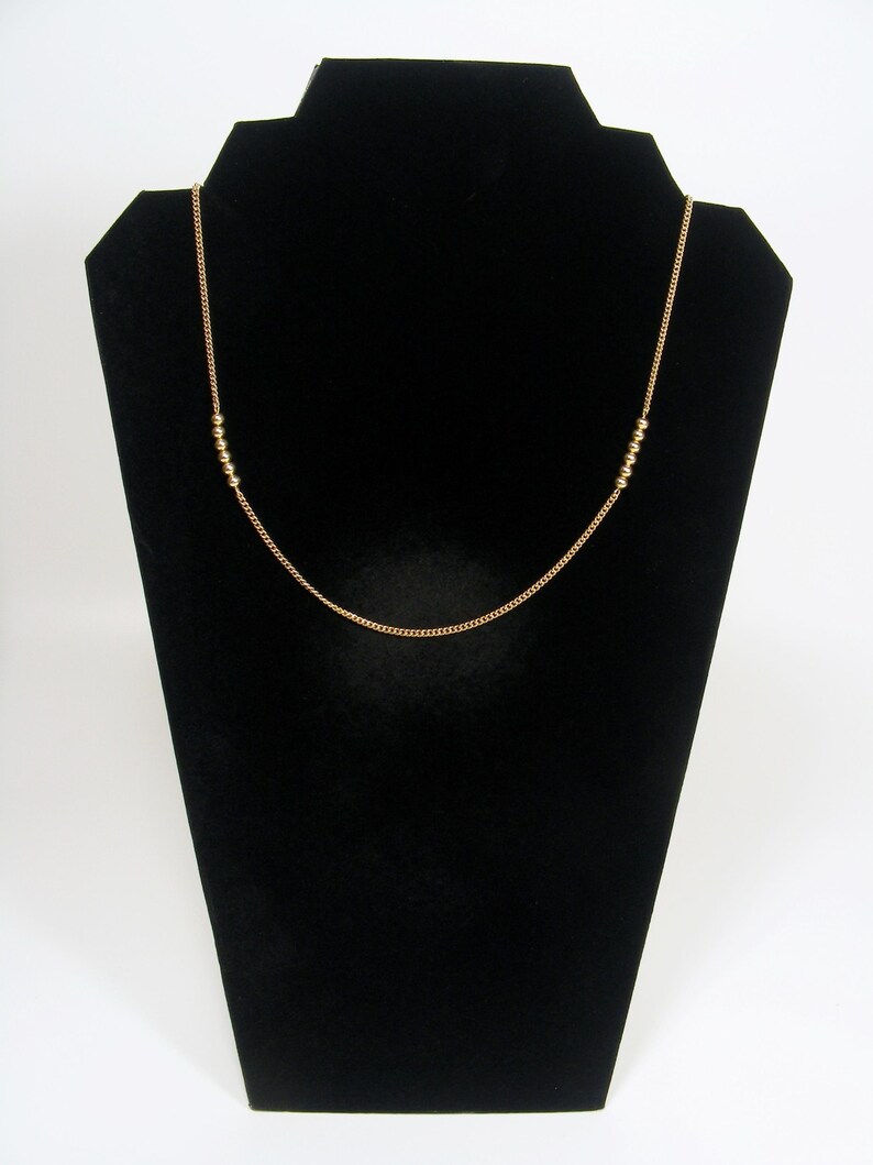 May include: A gold-coloured necklace on a black jewellery stand. The necklace has a fine chain with small, round beads spaced along it. The design is simple and elegant, suitable for everyday wear, measuring approximately 45 cm in length.