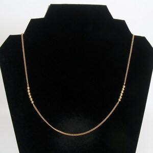 May include: A gold-coloured necklace featuring a fine chain and small, round beads. The necklace is presented on a black jewellery stand, highlighting its simple elegance. The beads are evenly spaced along the chain.