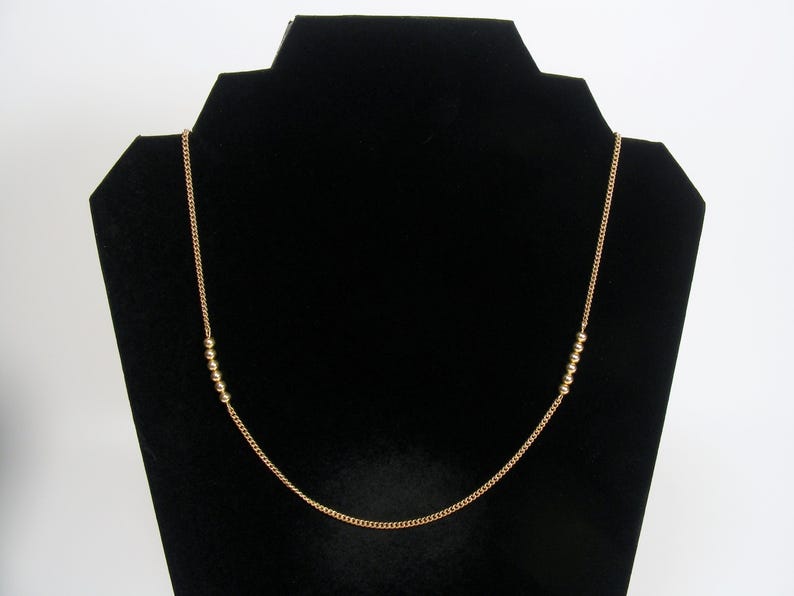 May include: A gold-coloured necklace featuring a fine chain and small, round beads. The necklace is presented on a black jewellery stand, highlighting its simple elegance. The beads are evenly spaced along the chain.