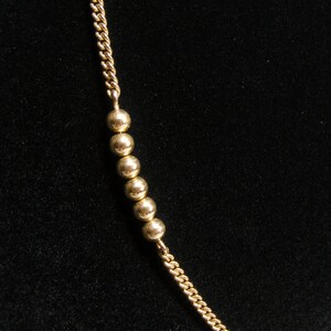May include: A gold-coloured necklace featuring a chain and a series of small, round beads. The beads are evenly spaced along the chain. The background is a solid black colour.