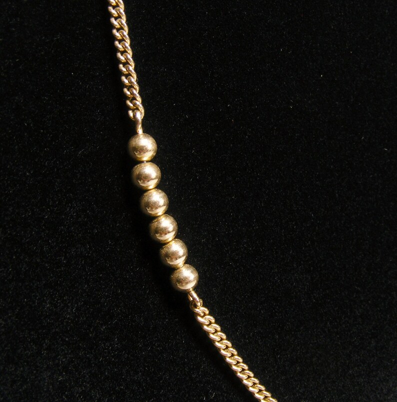 May include: A gold-coloured necklace featuring a chain and a series of small, round beads. The beads are evenly spaced along the chain. The background is a solid black colour.
