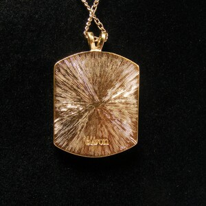 May include: Gold-coloured pendant necklace with a rectangular pendant. The pendant features a textured, sunburst design and the word "Avon" is inscribed at the bottom. The necklace is on a black background.