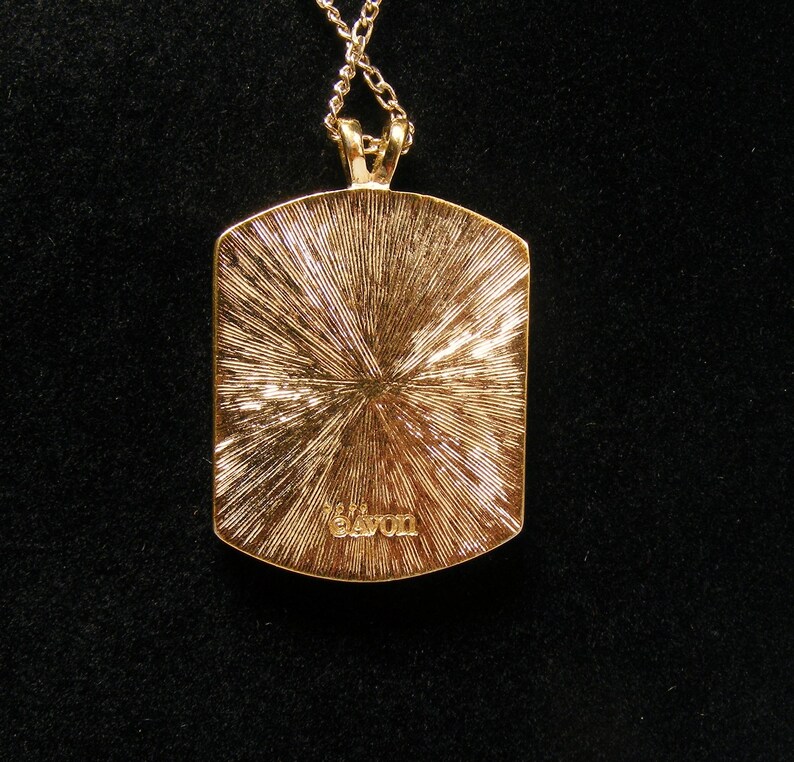 May include: Gold-coloured pendant necklace with a rectangular pendant. The pendant features a textured, sunburst design and the word "Avon" is inscribed at the bottom. The necklace is on a black background.