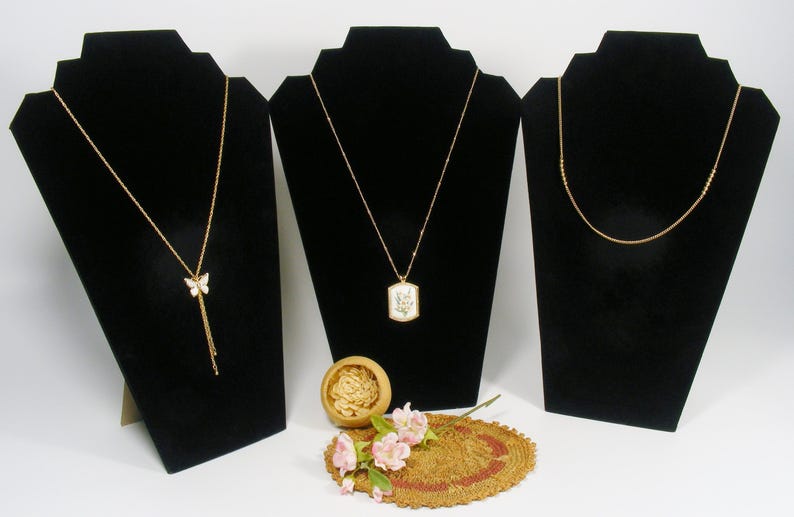 May include: Three gold necklaces displayed on black velvet busts. One has a butterfly pendant with dangling accents, another has a rectangular pendant with floral details, and the third has a simple beaded design. A small wooden bowl and floral arrangement are in the foreground.