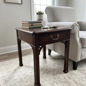 May include: A square dark wood end table featuring a single drawer with a decorative brass pull handle, straight fluted legs with carved corner brackets, and a smooth top surface holding a stack of three books, a small potted succulent, and a magnifying glass.