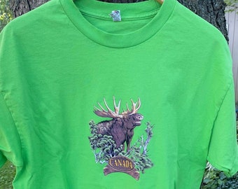 May include: A bright green t-shirt with a graphic of a moose in a forest setting. The moose is brown and has large antlers. The graphic also includes the word "CANADA" in a banner.