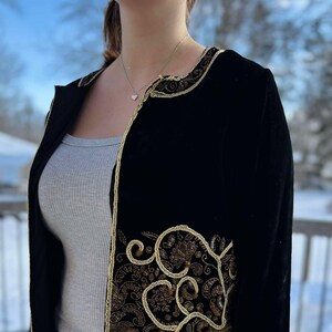 Peut inclure: Une veste en velours noir avec des broderies dorées. La broderie présente un motif floral avec des détails complexes. La veste a un col et des manches longues.
