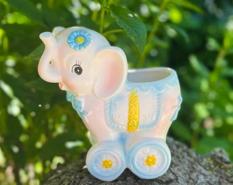 May include: A white ceramic elephant planter with blue and yellow details. The elephant has a flower on its head and wheels on its feet. It is sitting on a tree stump.