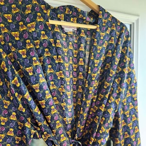 May include: A navy blue robe with a repeating pattern of yellow teddy bears and purple paisley shapes. The robe has long sleeves and a tie closure at the waist. The robe is hanging on a wooden hanger.