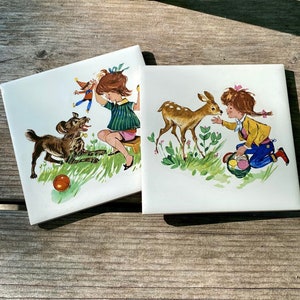 May include: Two vintage ceramic tiles with colorful illustrations. The first tile shows a girl playing with a dog and a ball. The second tile shows a girl with a basket of flowers and a fawn.
