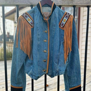 May include: A blue denim jacket with brown suede fringe and beaded accents on the shoulders. The jacket has a black velvet collar and brown suede trim around the buttons and cuffs.