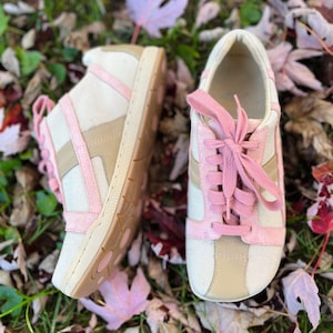 K&ouml;nnte beinhalten: Ein Paar wei&szlig;e Canvas-Sneaker mit rosa Akzenten und Schn&uuml;rsenkeln. Die Schuhe liegen auf einem Bett aus Herbstlaub.