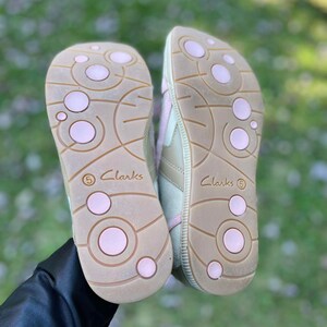 K&ouml;nnte beinhalten: Ein Paar beigefarbene Canvas-Sneaker mit rosa Akzenten. Die Schuhe haben eine strukturierte Sohle mit rosa Kreisen und das Wort "Clarks" ist an der Seite aufgedruckt.
