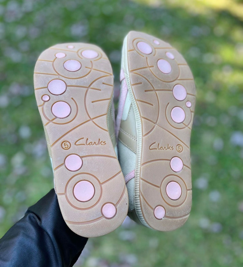 K&ouml;nnte beinhalten: Ein Paar beigefarbene Canvas-Sneaker mit rosa Akzenten. Die Schuhe haben eine strukturierte Sohle mit rosa Kreisen und das Wort "Clarks" ist an der Seite aufgedruckt.
