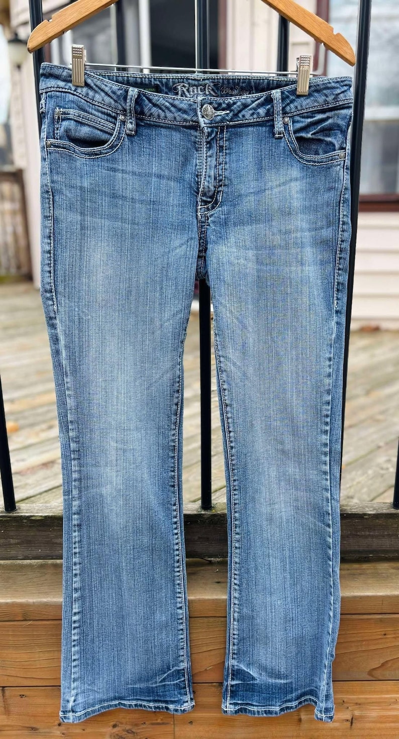 Puede incluir: Un par de pantalones vaqueros azules de mezclilla con pierna acampanada. Los vaqueros tienen un lavado claro y est&aacute;n colgados en una percha de madera.