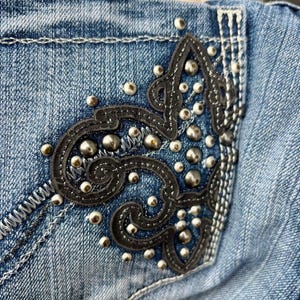 Puede incluir: Vaqueros azules de denim con un dise&ntilde;o de cuero negro y tachuelas plateadas en el bolsillo trasero.