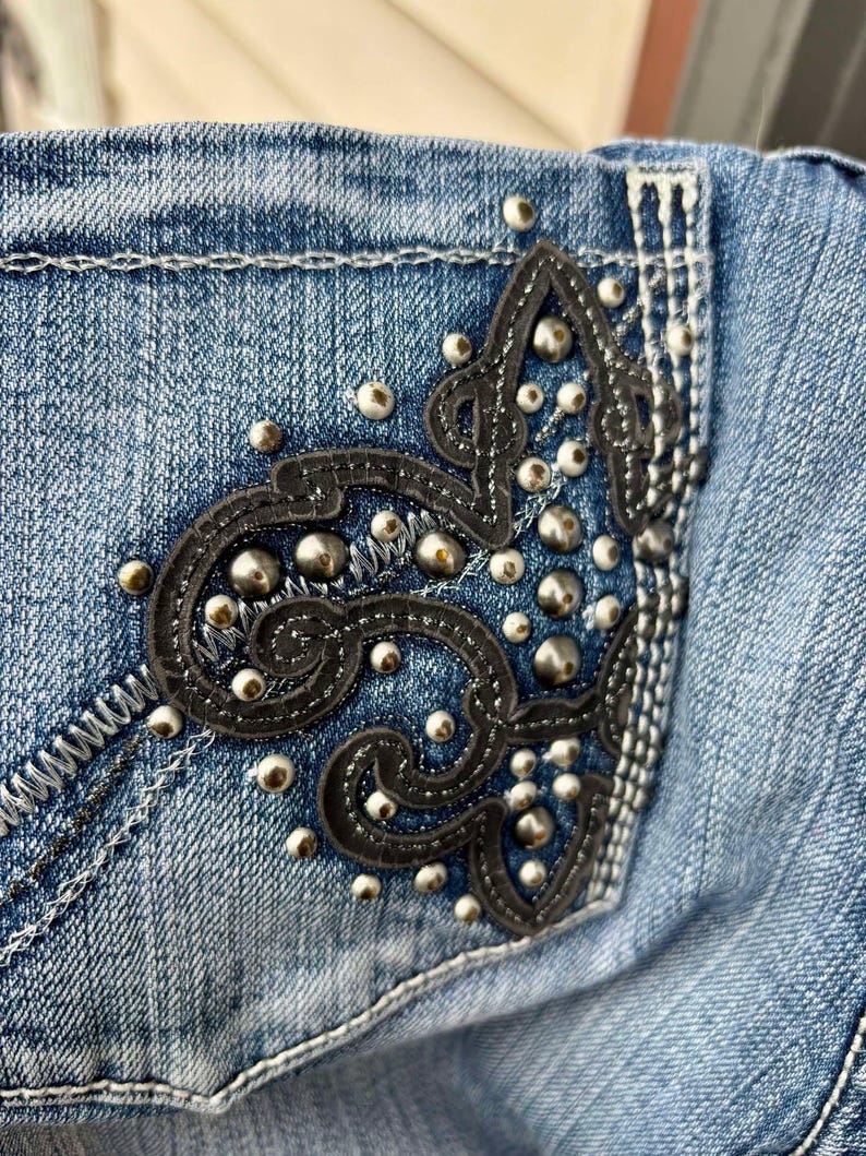 Puede incluir: Vaqueros azules de denim con un dise&ntilde;o de cuero negro y tachuelas plateadas en el bolsillo trasero.