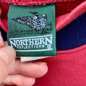 May include: A green tag with a loon bird logo and the text "Northern Reflections" in white lettering. The tag is attached to a red sweatshirt with a navy blue trim.