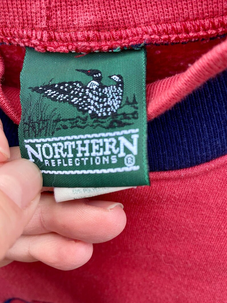 May include: A green tag with a loon bird logo and the text "Northern Reflections" in white lettering. The tag is attached to a red sweatshirt with a navy blue trim.
