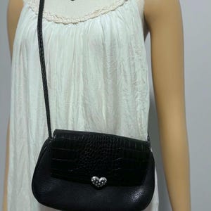 May include: Black crossbody bag with a textured flap and a silver heart-shaped clasp. The bag has a braided strap and is worn over a white dress. The bag's design includes a crocodile-like pattern.
