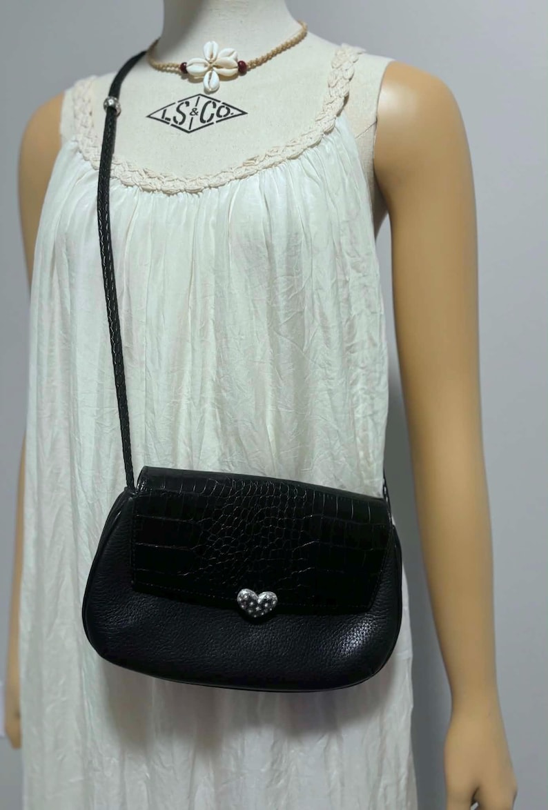 May include: Black crossbody bag with a textured flap and a silver heart-shaped clasp. The bag has a braided strap and is worn over a white dress. The bag's design includes a crocodile-like pattern.