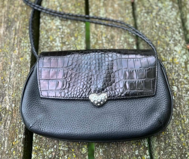 May include: Black crossbody bag with a textured leather body and a glossy, faux-crocodile flap. The flap is secured with a silver heart-shaped clasp. The bag has a braided strap and is set against a natural wood background.