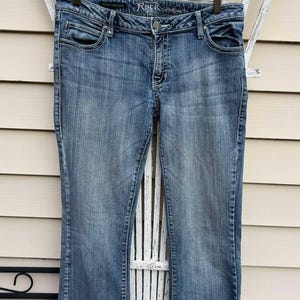 Puede incluir: Un par de pantalones vaqueros azules de mezclilla con pierna acampanada. Los vaqueros tienen un lavado oscuro y est&aacute;n colgados en una percha de madera. La marca "Rock & Republic" es visible en la cintura.