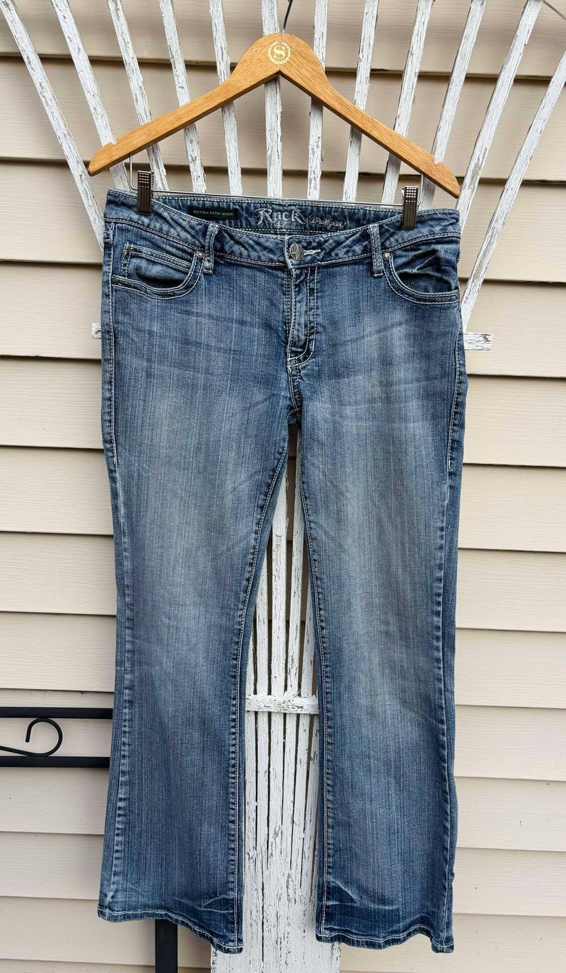 Puede incluir: Un par de pantalones vaqueros azules de mezclilla con pierna acampanada. Los vaqueros tienen un lavado oscuro y est&aacute;n colgados en una percha de madera. La marca "Rock & Republic" es visible en la cintura.