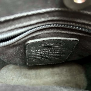 May include: Interior view of a black handbag. The image shows the inside lining, a zipper, and a small black leather tag with the Brighton logo and text. The bag appears to be made of a soft, dark material.