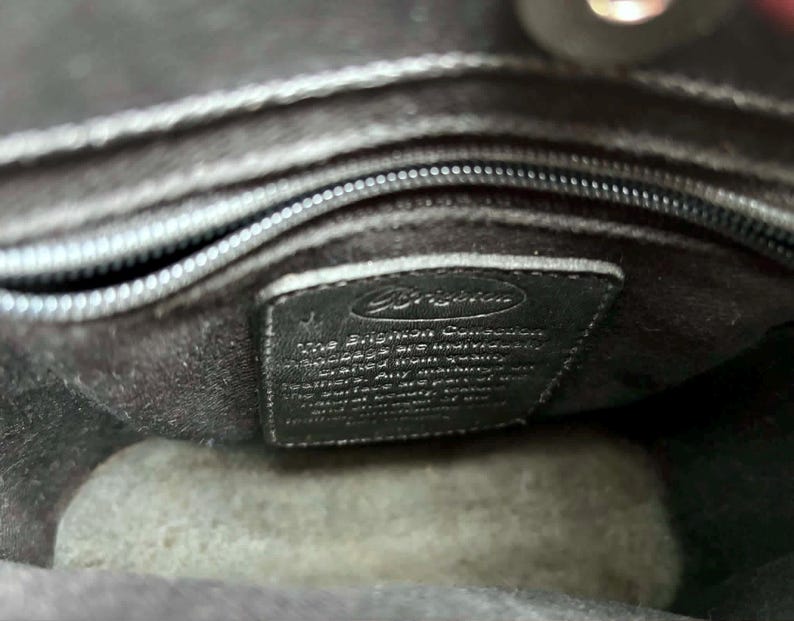 May include: Interior view of a black handbag. The image shows the inside lining, a zipper, and a small black leather tag with the Brighton logo and text. The bag appears to be made of a soft, dark material.