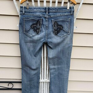 Puede incluir: Un par de pantalones vaqueros azules con bolsillos traseros adornados. Los adornos son negros y presentan un dise&ntilde;o en forma de diamante con pedrer&iacute;a.