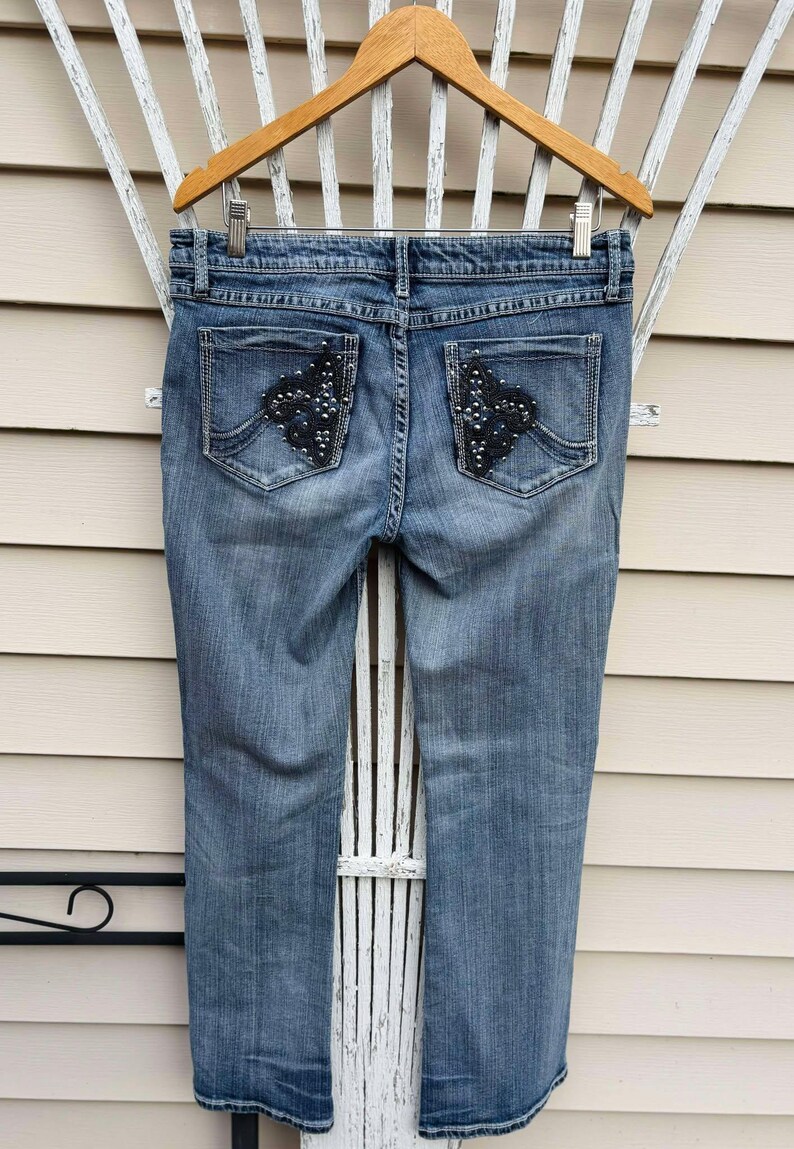 Puede incluir: Un par de pantalones vaqueros azules con bolsillos traseros adornados. Los adornos son negros y presentan un dise&ntilde;o en forma de diamante con pedrer&iacute;a.